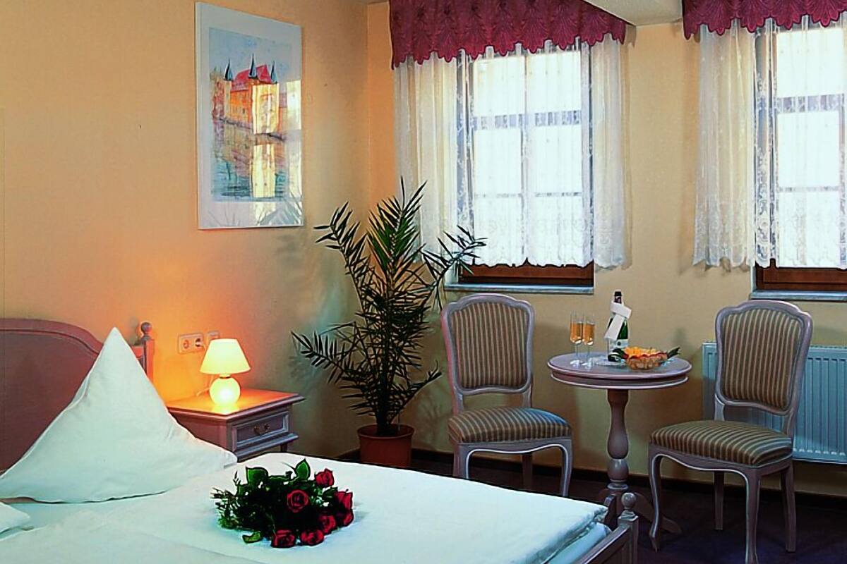 Hotelkamer met een bed, nachtkastje met een lamp, een potplant, twee fauteuils, een tafeltje met een fles en glazen, en een raam met gordijnen.