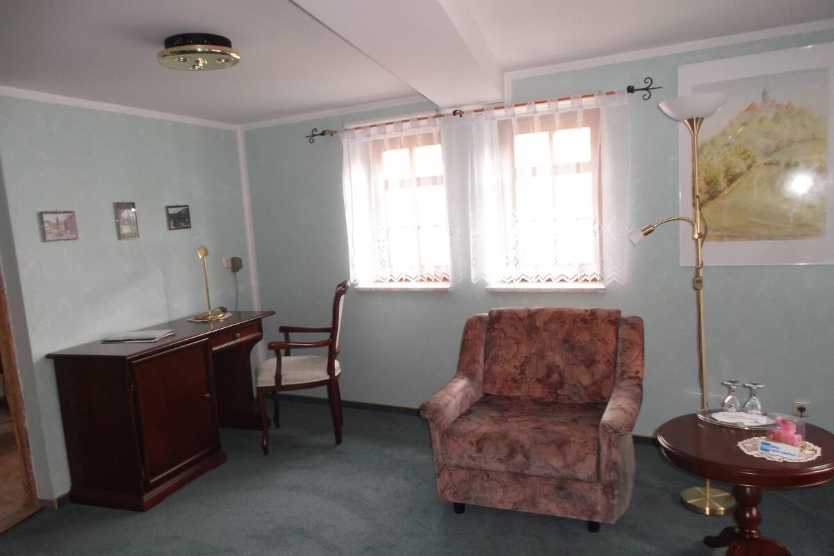 Een kamer met een bureau, een stoel, een bank, een tafel met een lamp en een raam met gordijnen.