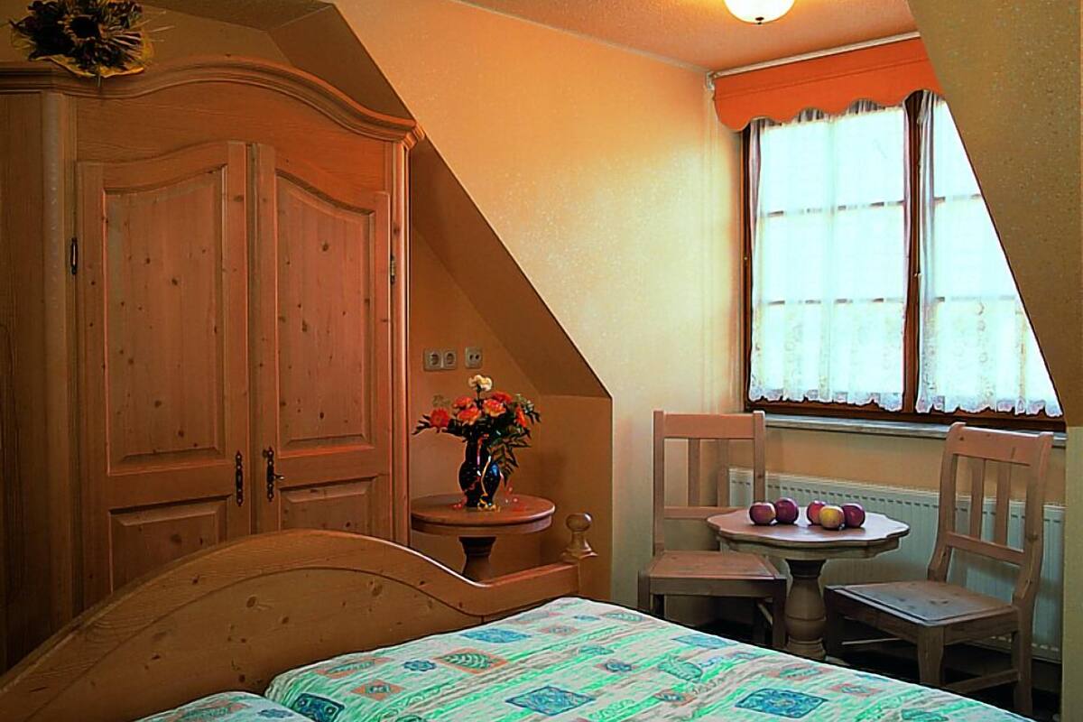 Een slaapkamer met een schuine kap, een houten garderobe, twee eenpersoonsbedden, een klein tafeltje met een stoel en een raam met gordijnen.