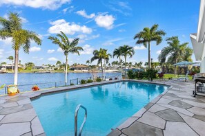 A swimming pool with a clear view of the water, surrounded by palm trees and a building in the background. - Centrally Located, Waterfront, Modern Home with lots of amenities (Cape Coral)