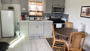 A modern kitchen with grey cabinets, a stainless steel stove, and a microwave. There's a dining area with a wooden table and chairs, and a welcome mat on the floor. - New Private Studio - UCR - LLU (Riverside)