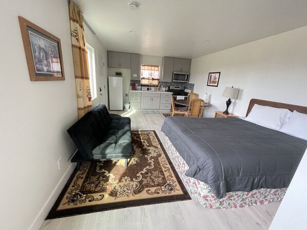 A compact bedroom with a bed, a sofa, and a kitchenette in the background.