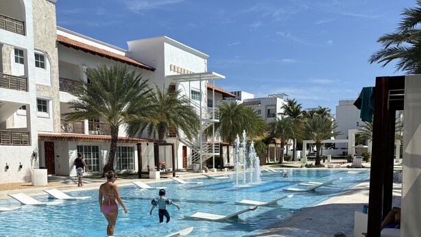 A swimming pool area with a clear sky, palm trees, and a building with balconies. - Confy apart CAP CANA (Punta Cana)