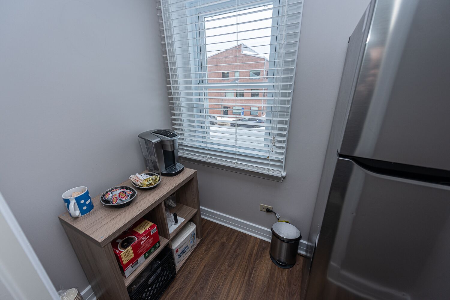 A small kitchenette with a coffee maker, a mug, a plate with cookies, and a trash can.