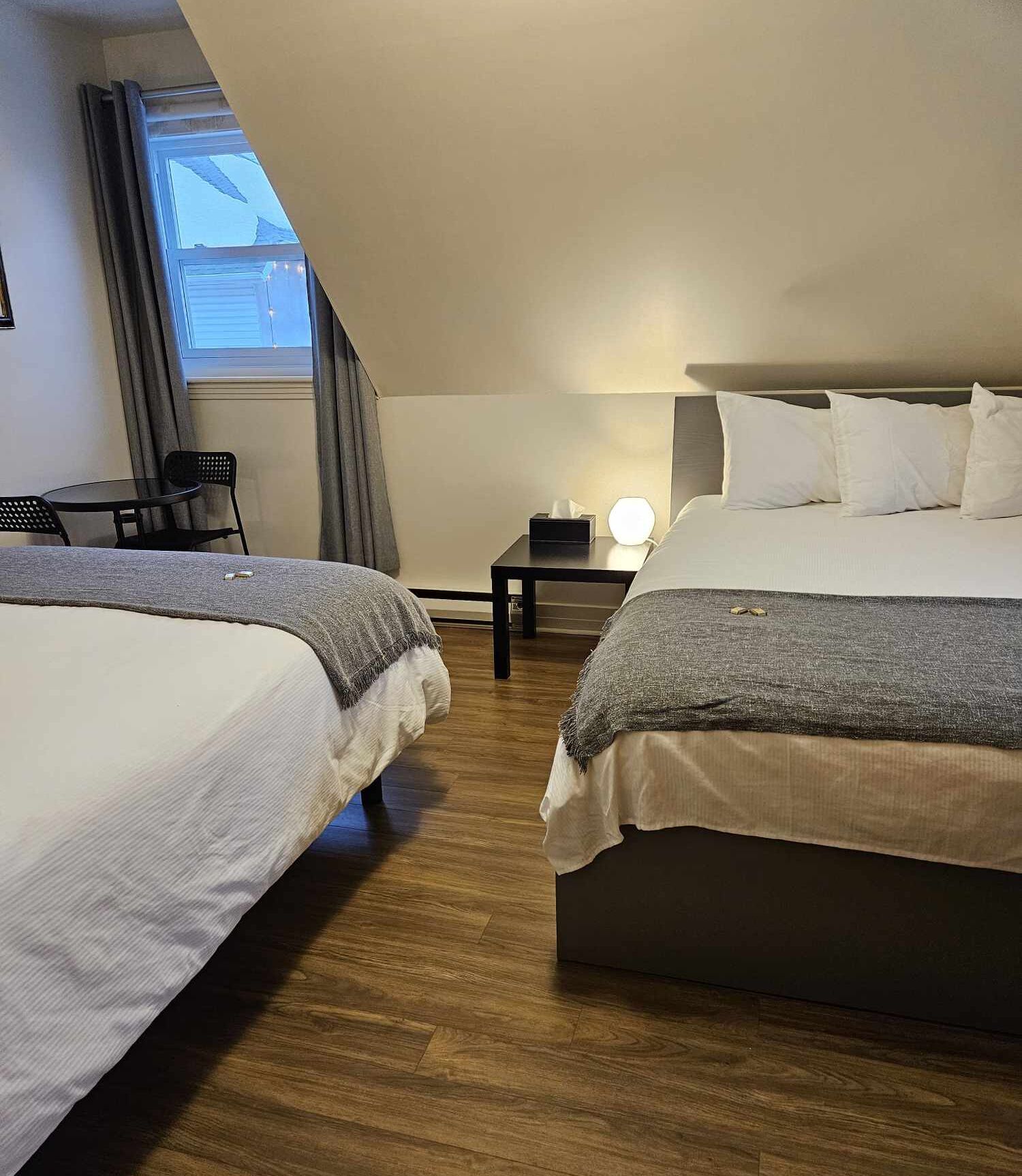 A hotel room with two beds, a small table, a chair, and a window with curtains.