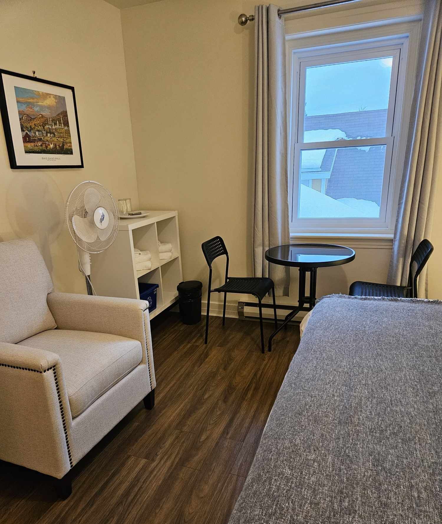 A hotel room with a bed, a chair, a small table, a fan, a shelf with towels, and a window with a view of buildings.