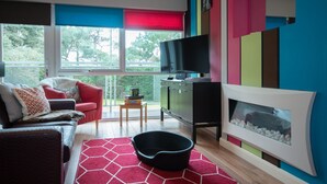 A modern living room with a sofa, a television, a fireplace, and a view of greenery outside. - Two bedroom apartment with balcony dog friendly  (Sandbanks)