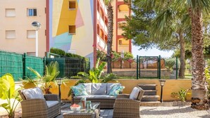 A sunlit outdoor seating area with wicker furniture, a glass-top table, and a view of a colorful building in the background.
