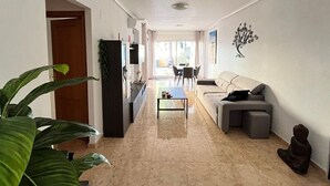 A spacious living room with a sofa, a coffee table, and a TV. There is a dining area with chairs and a table in the background.