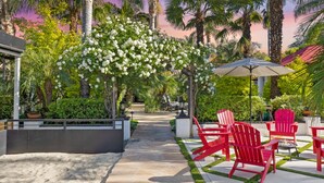 A tropical garden patio with red Adirondack chairs, a pathway, and a pergola with white flowers. - Erwin Estate (Woodland Hills)