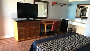 Habitación de hotel con escritorio de madera, televisor, silla de oficina roja, cama con colcha estampada y baño con espejo y lavamanos.
