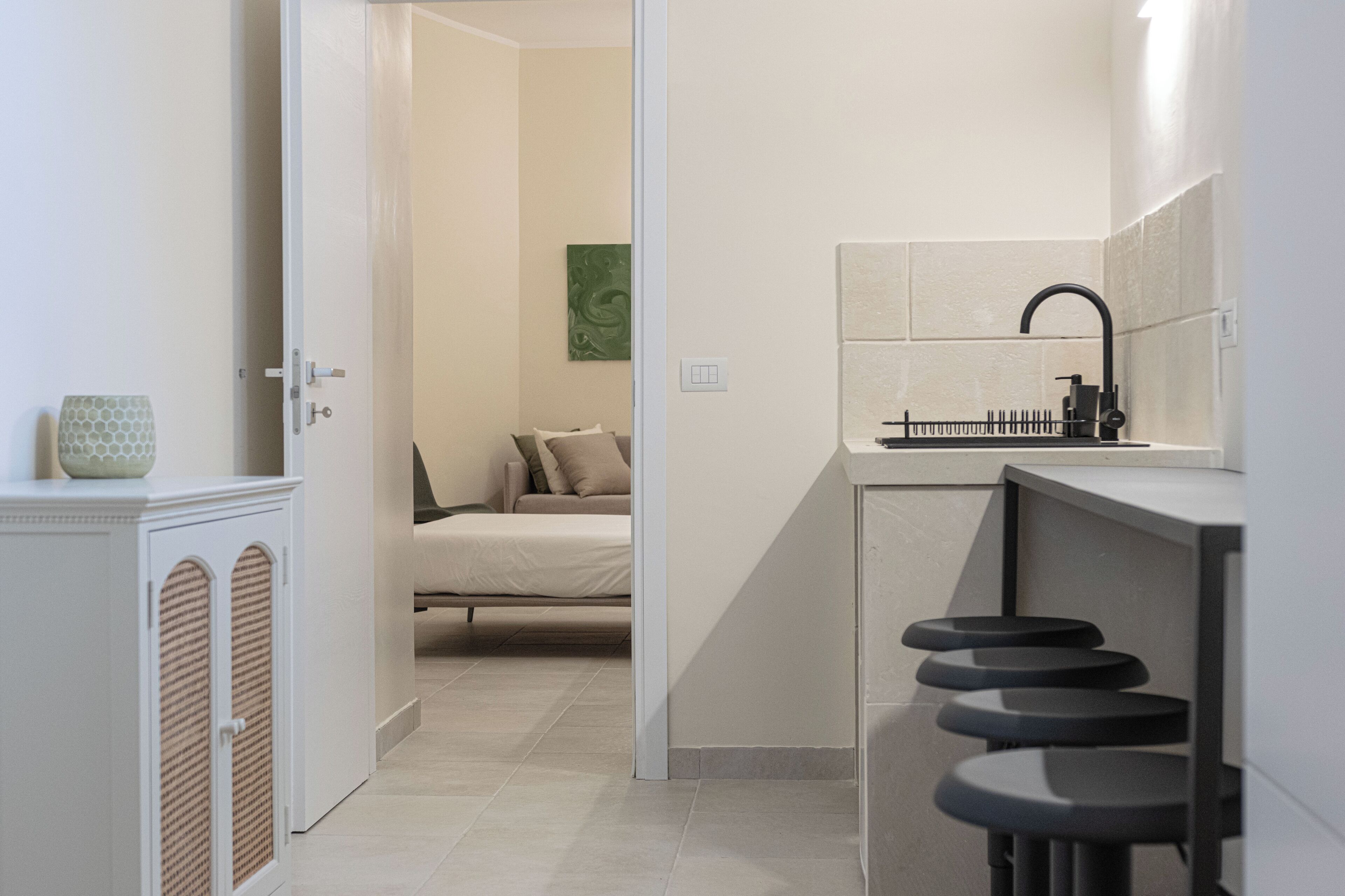 A modern kitchen with a black faucet, a small island with stools, and a door leading to a bedroom with a bed and pillows.