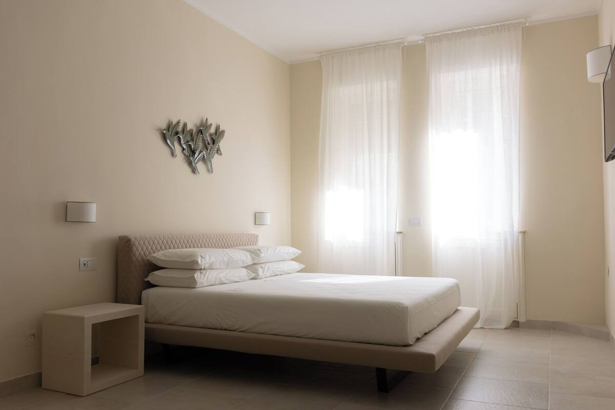 A bedroom with a bed, bedside tables, a wall-mounted lamp, and sheer curtains.