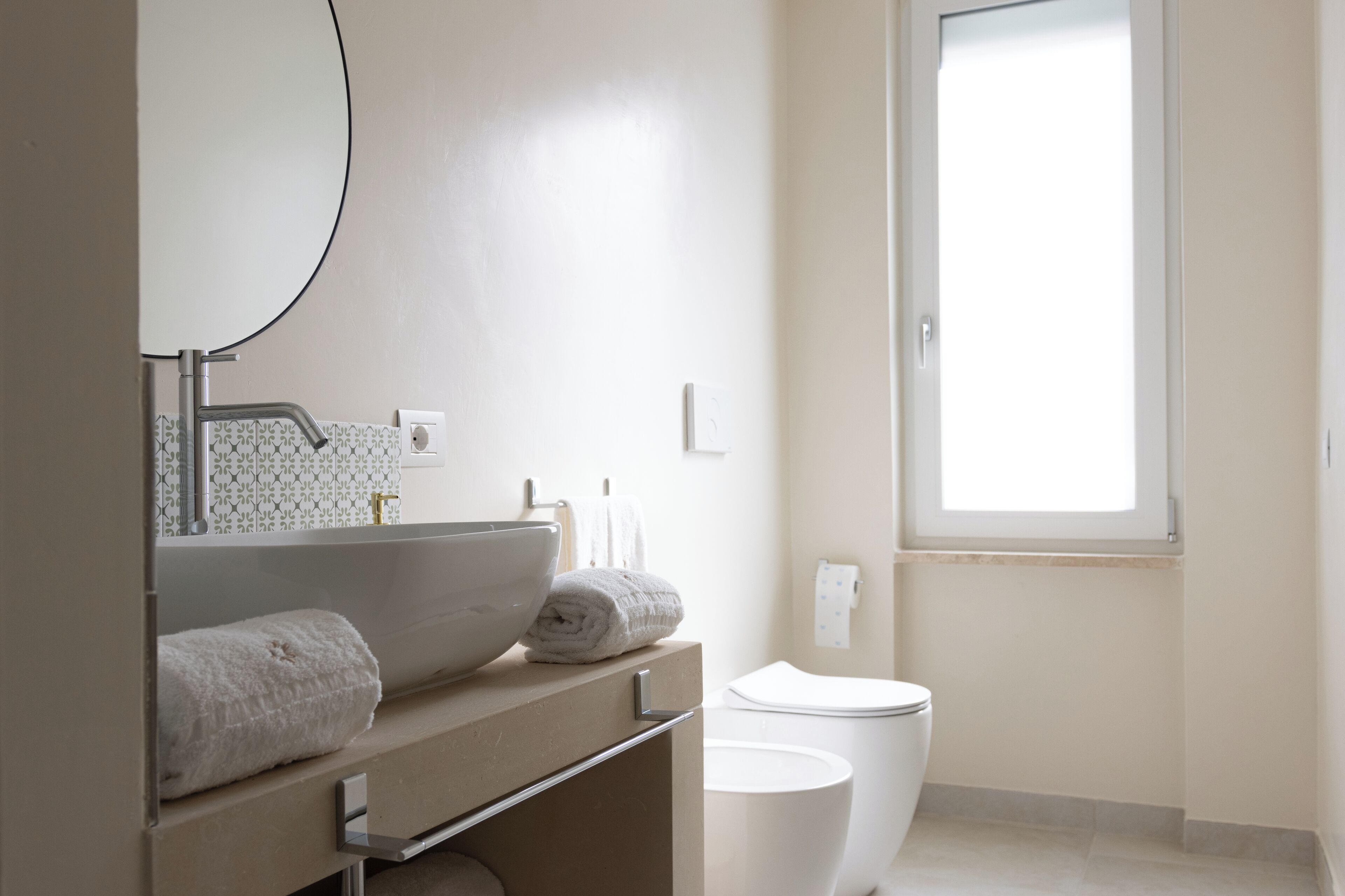 A minimalist bathroom with a large mirror, a white sink, a toilet, and a window providing natural light.