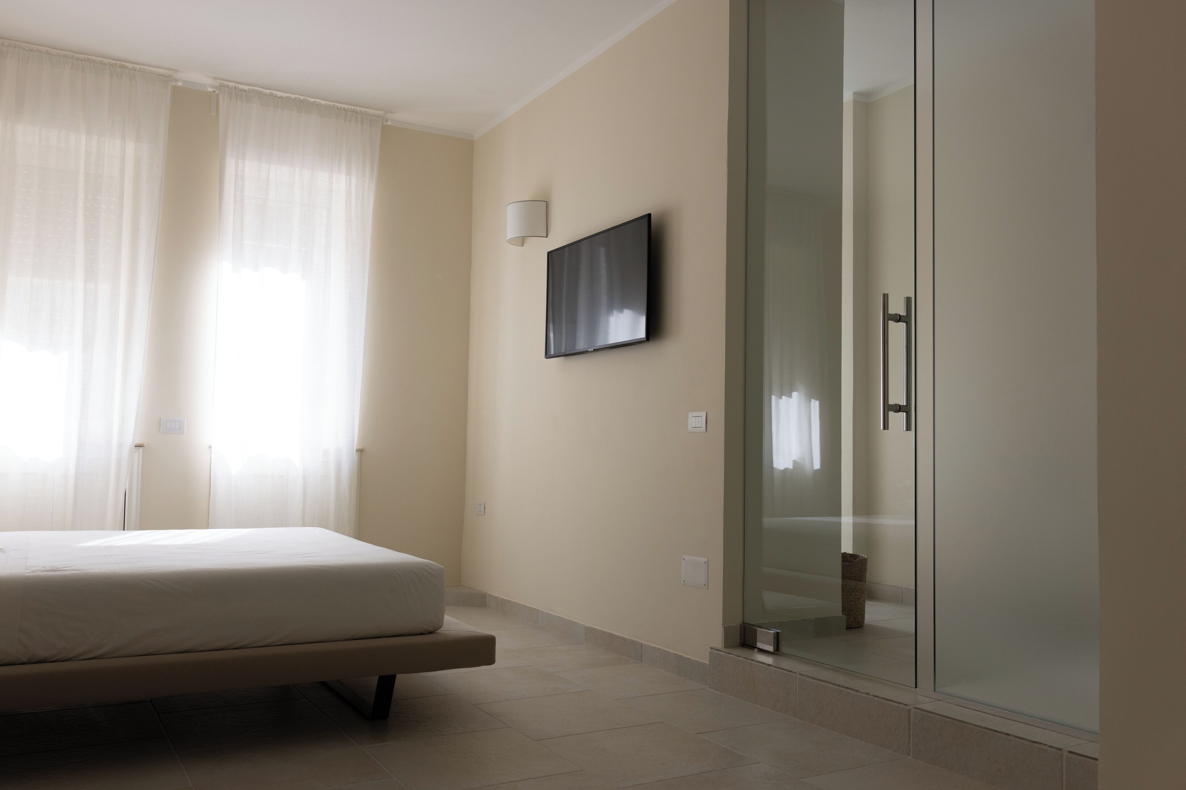 A bedroom with a bed, a glass shower enclosure, and a mounted flat-screen TV.