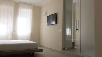 A bedroom with a bed, a glass shower enclosure, and a mounted flat-screen TV.
