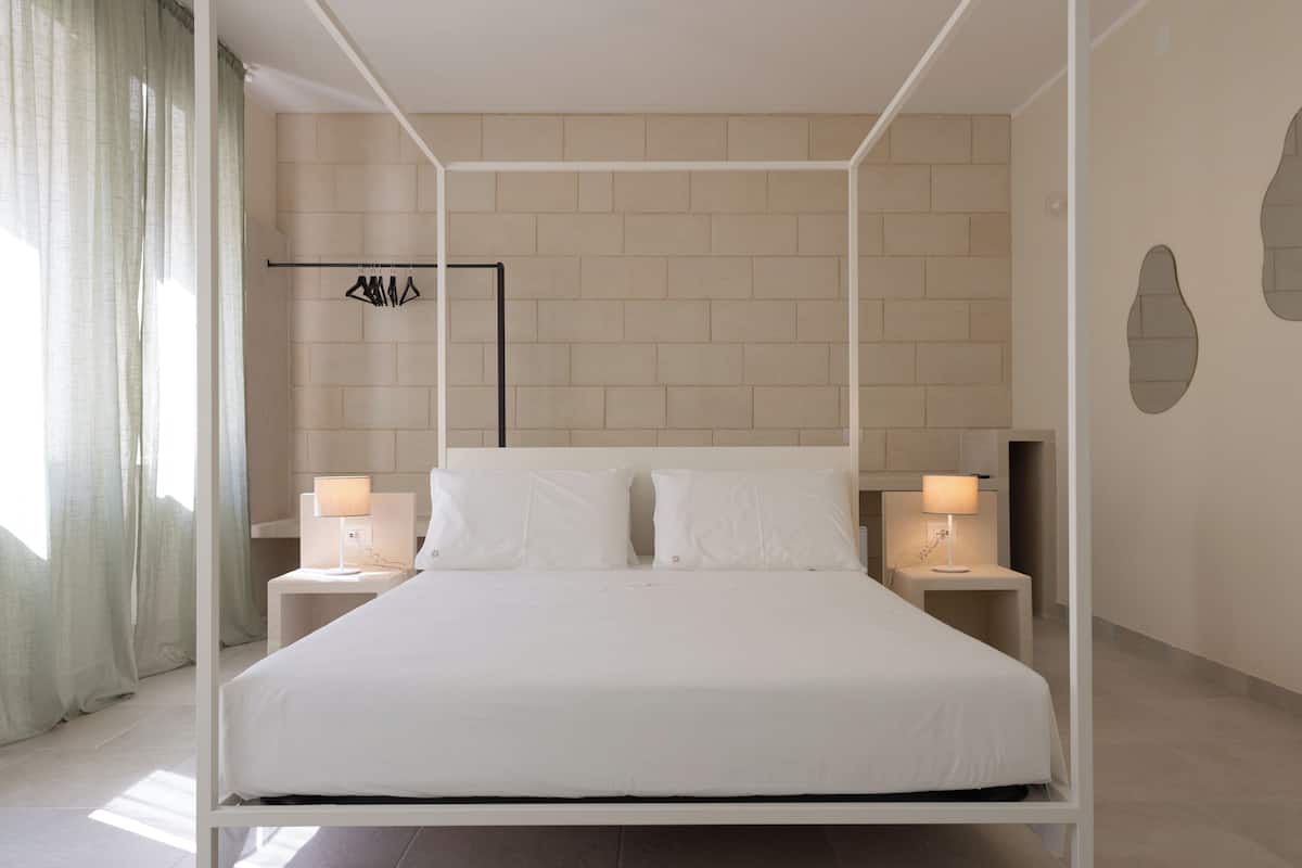 A modern bedroom with a four-poster bed, two bedside tables with lamps, a wardrobe, and a brick-patterned wall.