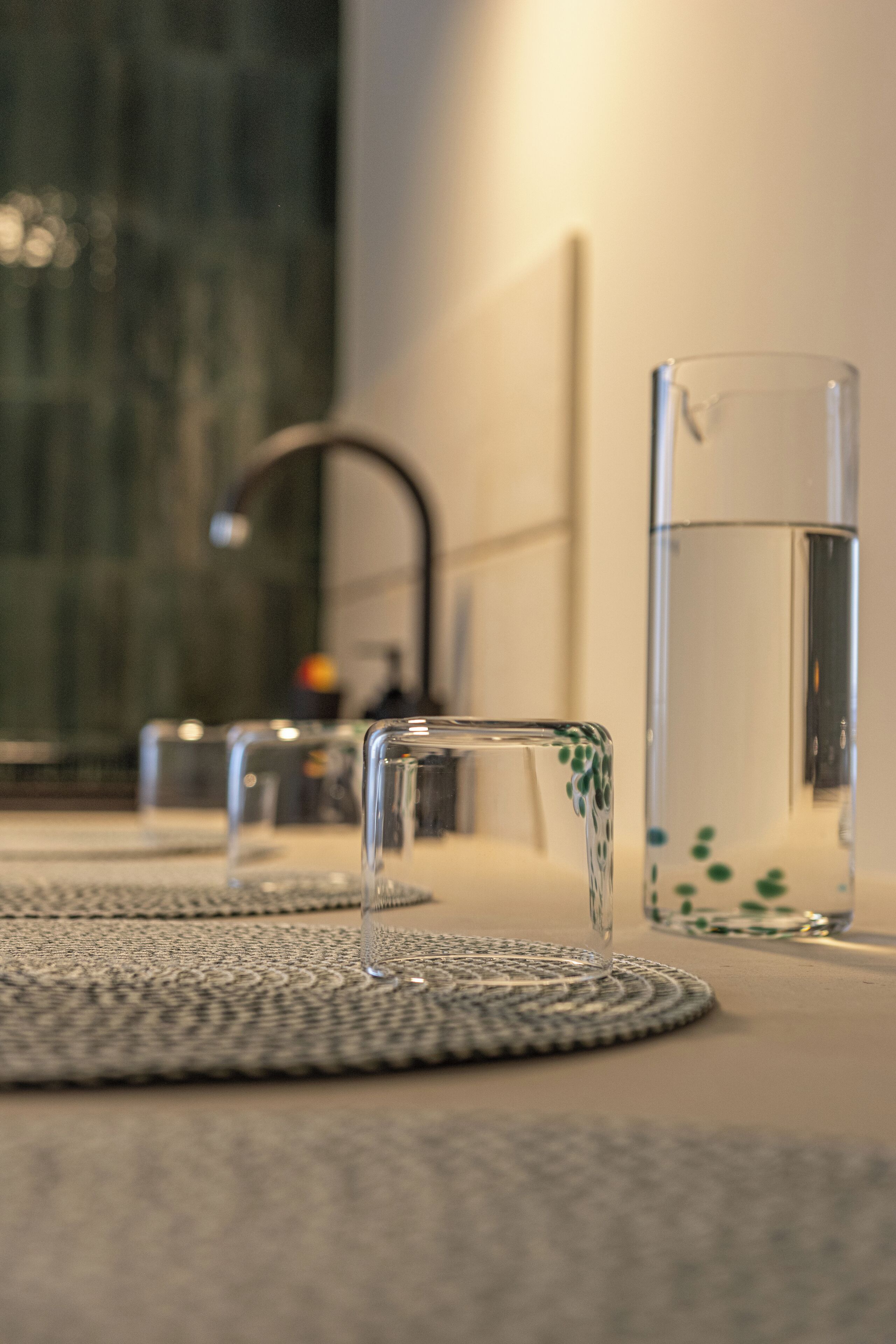 Een kamer met een transparante glazen tafel, een hoge glazen vaas en een klein glaasje op een geweven onderzetter.