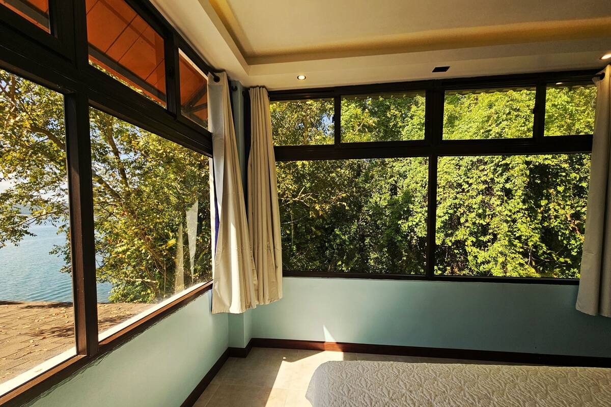 Ein Zimmer mit großem Fenster, einem Bett und Blick auf Bäume und Wasser.