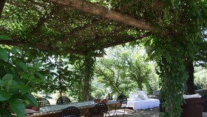 An outdoor dining area with a pergola, a table, chairs, and a bed under a tree.