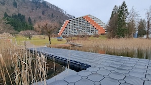 Ein sechseckig gemustertes Dock, das über ruhiges Wasser führt, umgeben von Schilf und Bäumen, mit einem modernen Gebäude im Hintergrund.