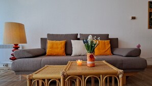 Ein Wohnzimmer mit grauem Sofa, orangefarbenen und weißen Kissen, einem kleinen Tisch mit Kerze und einer Blumenvase.