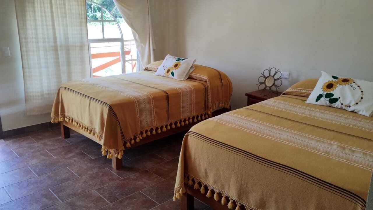 A room with two beds, each with a brown bedspread and matching pillows. There is a window with curtains, a small table with a decorative item, and a tiled floor.