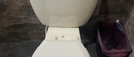 Een toilet met deksel en bedieningshendel, een prullenbak en een betegelde vloer.