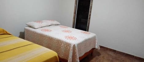 Una habitación pequeña con una cama individual, una pared blanca lisa y piso de baldosas.