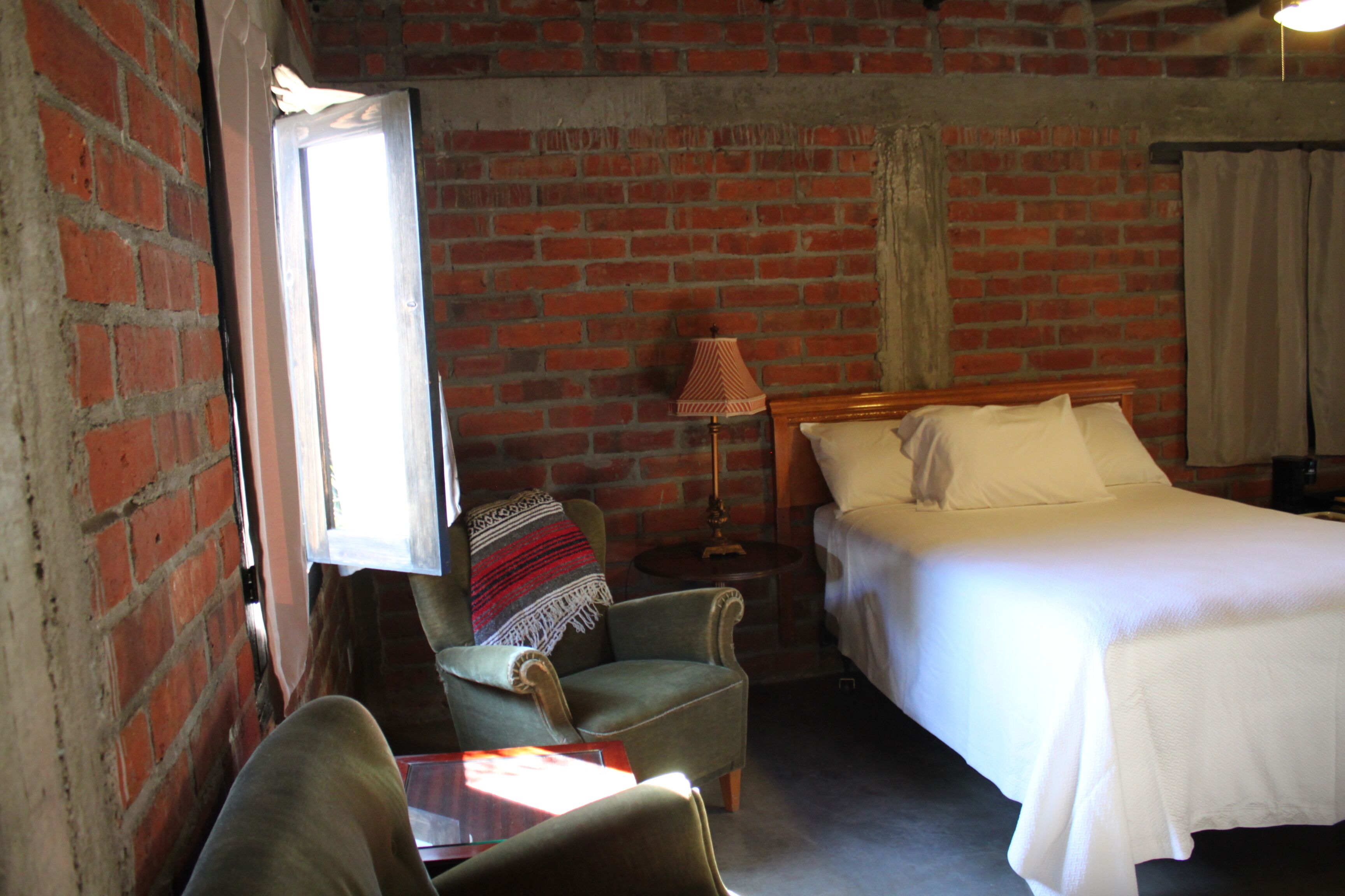 Une pièce avec un mur en briques, un lit recouvert de draps blancs, une chaise avec une couverture, une lampe et une table.