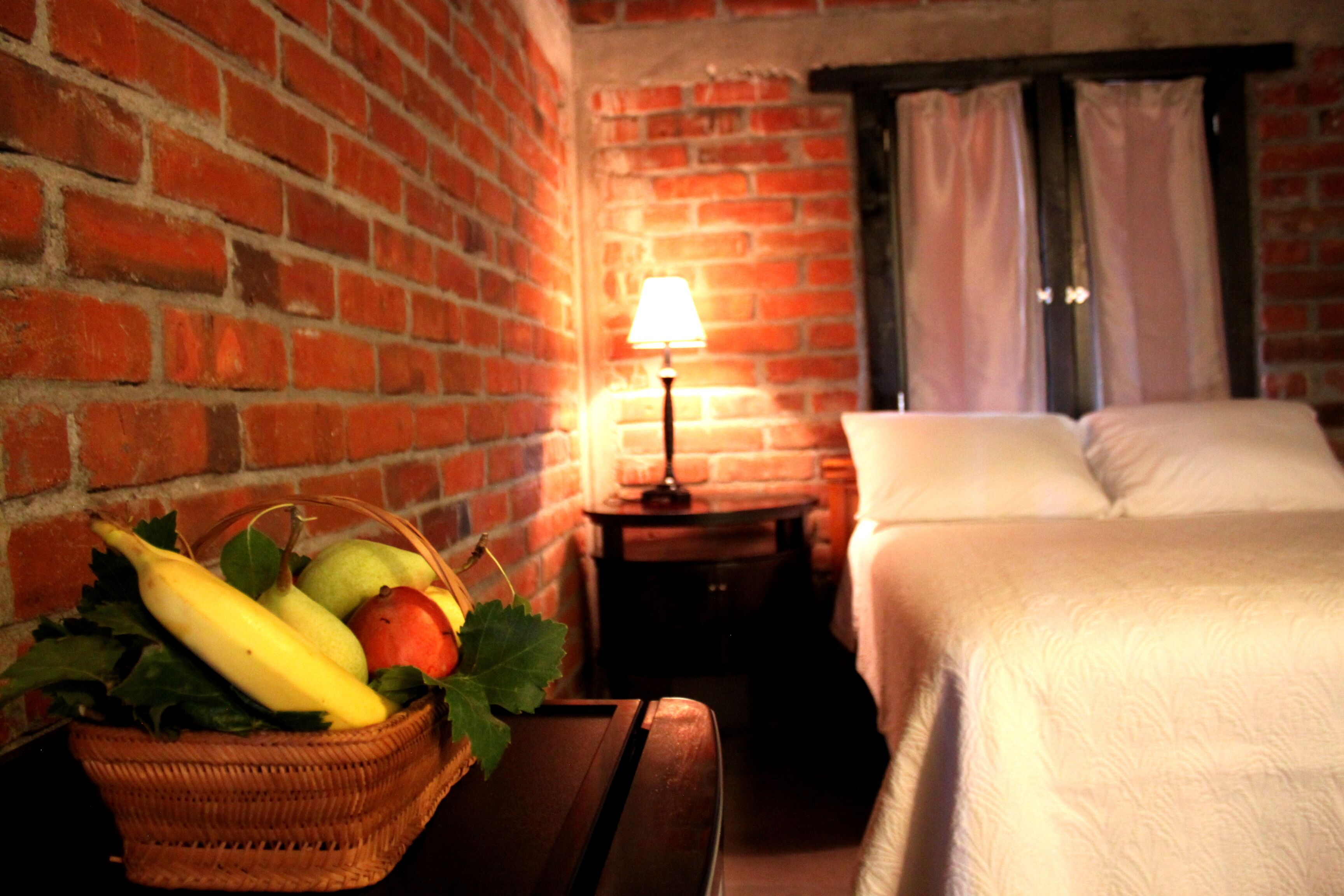 Ein Zimmer mit Backsteinwand, einem Bett mit weißer Bettwäsche, einem kleinen Tisch mit Lampe und einem Obstkorb.