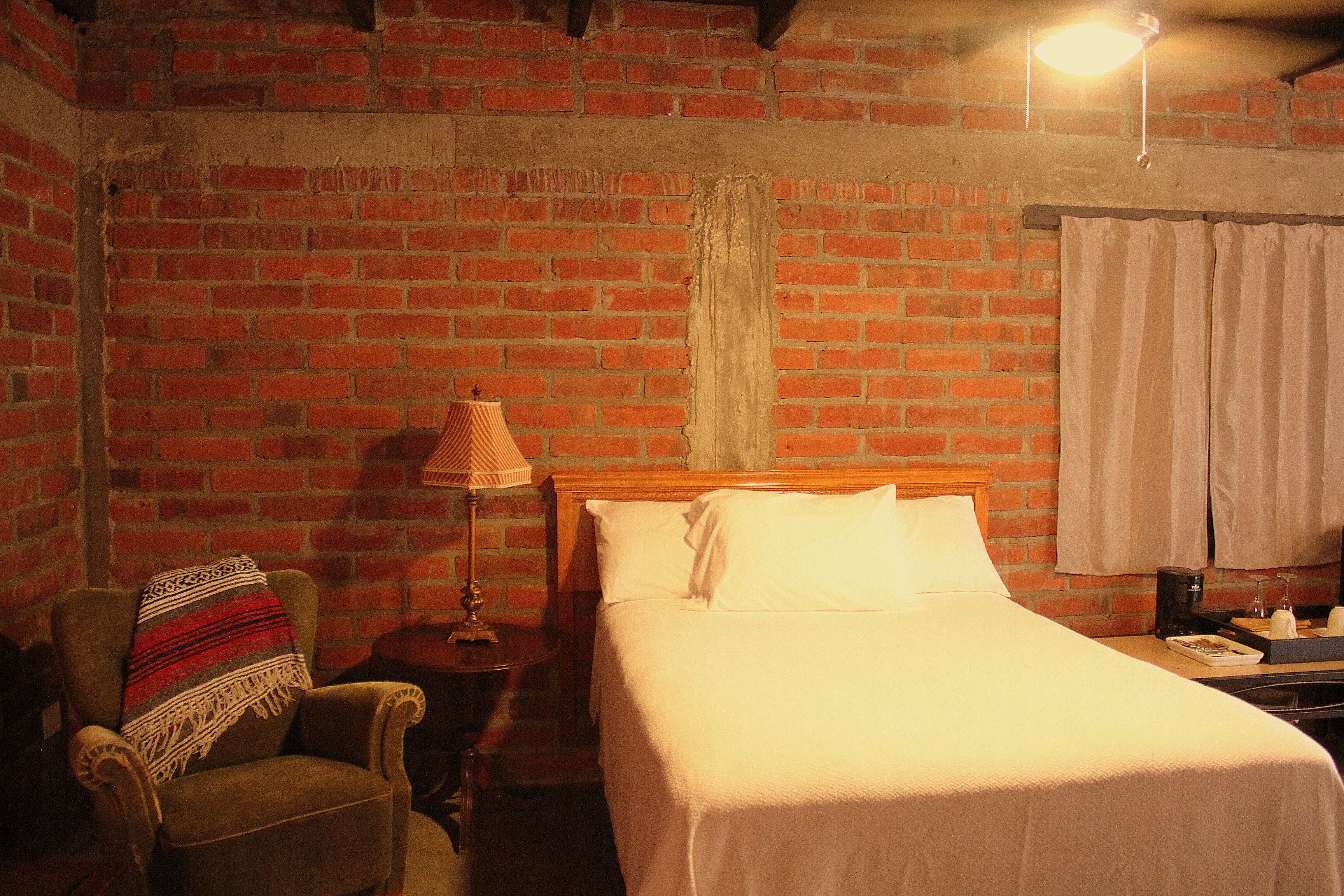 Quarto com parede de tijolos, cama de madeira, cadeira com manta, luminária e mesa de cabeceira com telefone e uma xícara.