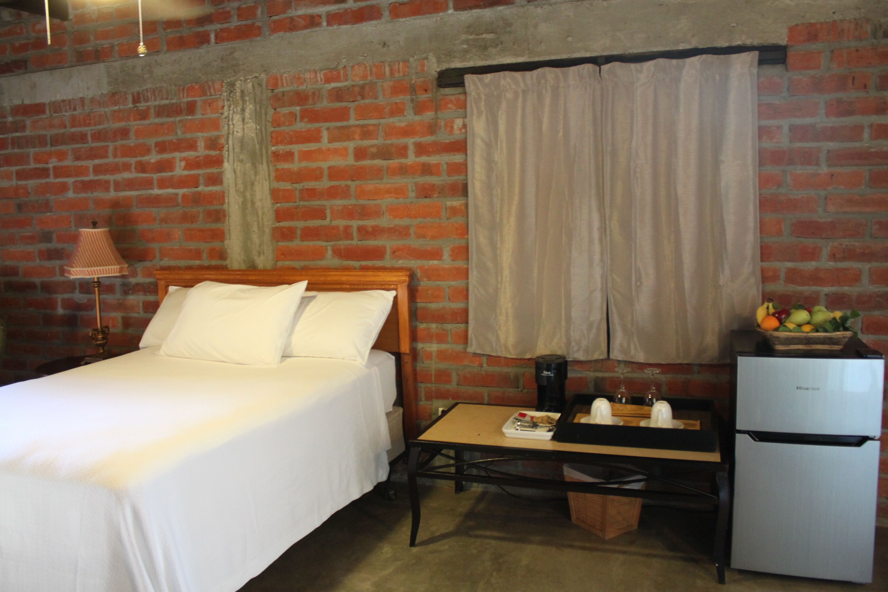 A room with a bed, a small table, a refrigerator, and a brick wall.