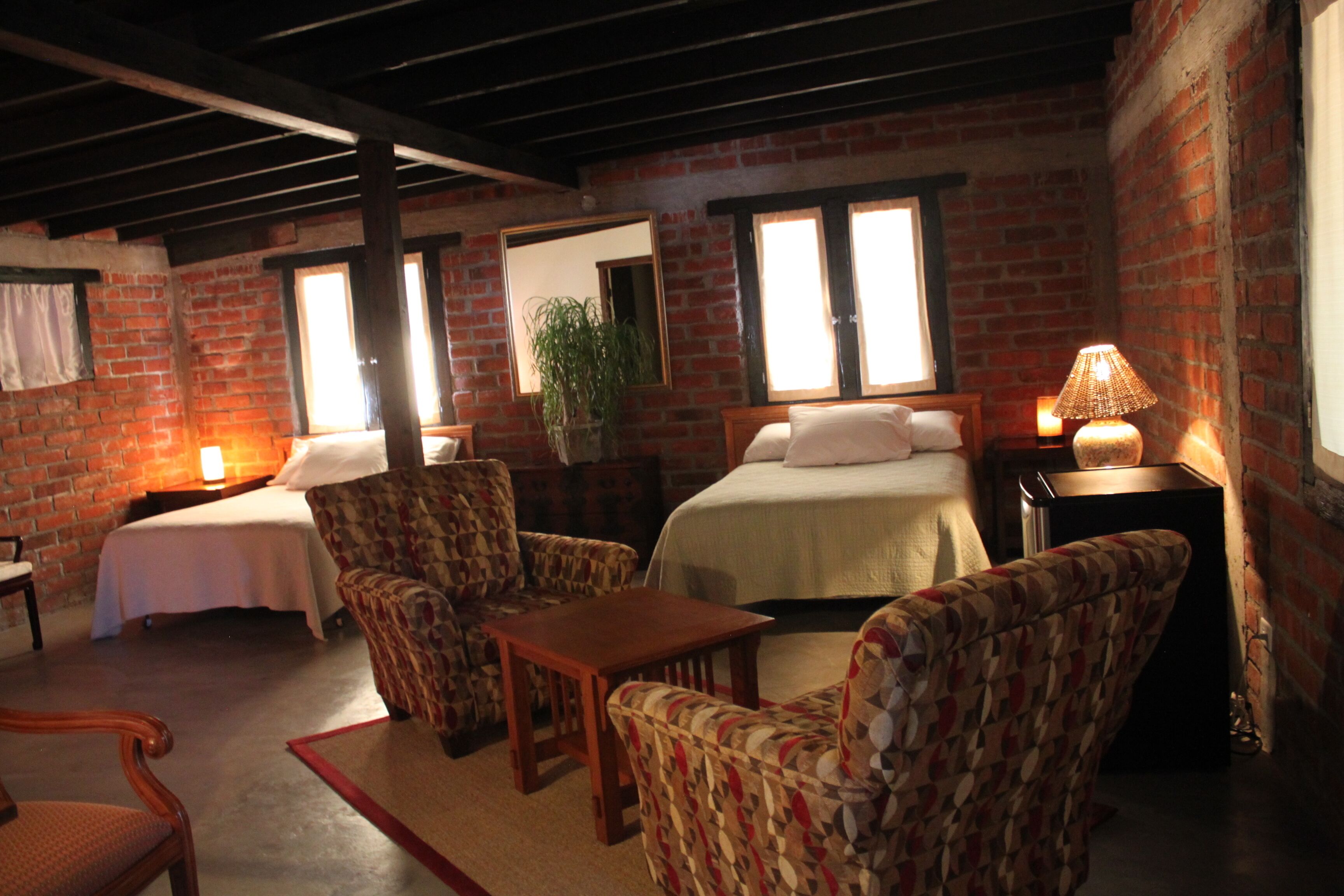 Una habitación con pared de ladrillo, una cama, dos sillas, una mesa y una lámpara.