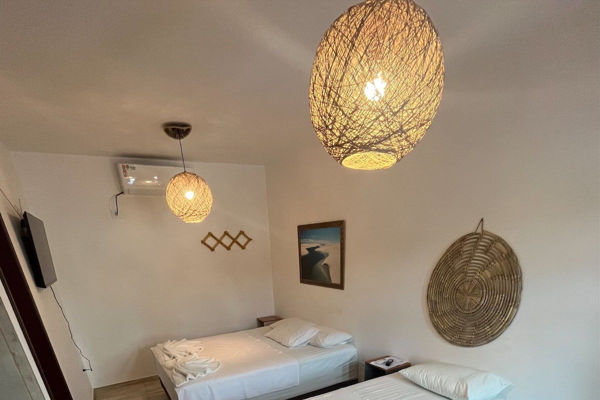 A hotel room with two beds, a small refrigerator, a wall-mounted air conditioner, a woven light fixture, and a wall art piece.