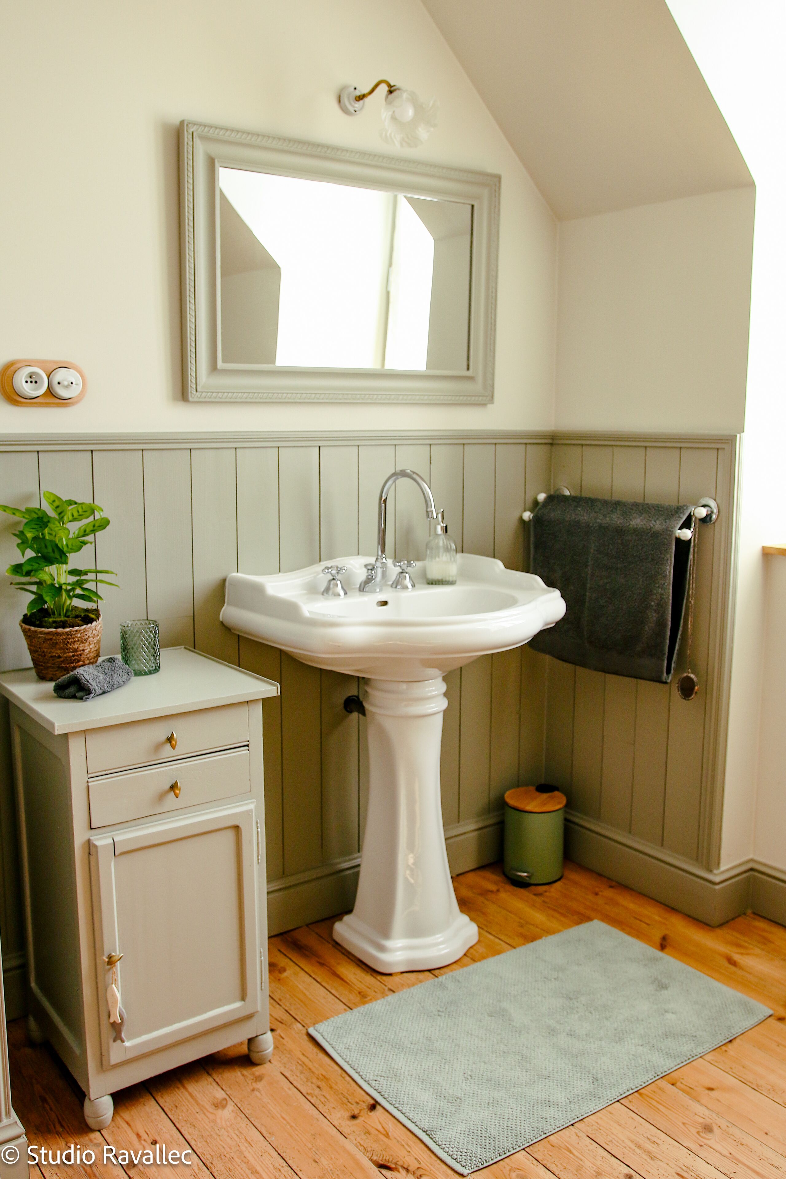 A bathroom with a pedestal sink, a mirror above it, a vanity with drawers, a potted plant, and a towel hanging on a rack.