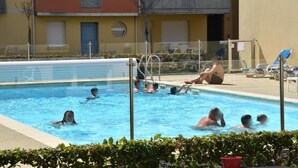 Ein Schwimmbecken, umgeben von einem Zaun, mit Liegestühlen und einem Gebäude im Hintergrund.