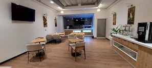 Un comedor moderno con mesas y sillas de madera, una cocina visible a través de una puerta abierta y un televisor de pantalla plana montado en la pared.