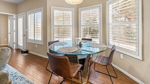 A dining area with a glass table, four chairs, and large windows with blinds.