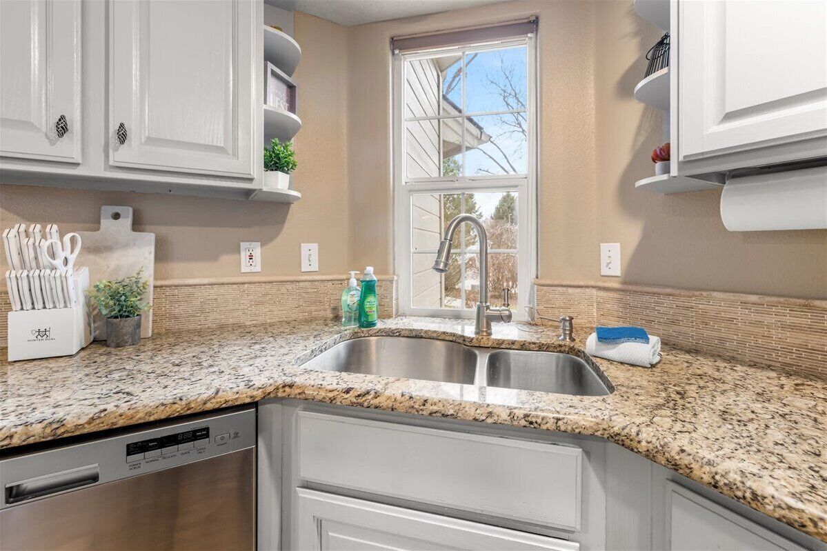 Una cocina con encimeras de granito, pileta de acero inoxidable, gabinetes blancos y una ventana con vista a árboles.