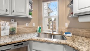 A kitchen with granite countertops, stainless steel sink, white cabinets, and a window with a view of trees.