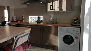 A compact kitchen with wooden cabinets, a built-in washing machine, a sink, and a dining area with a chair. - Joli T1 au Calme, Entre Collines et Littoral; Idéal Pour Explorer la Région! (Gémenos)