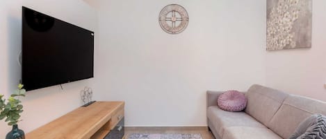 Ett vardagsrum med en platt-TV som är monterad på väggen, ett träbord, en mönstrad matta, en beige soffa med en kudde och en väggklocka.