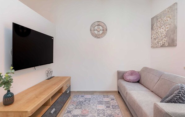 Una sala de estar con una televisión de pantalla plana montada en la pared, un mueble bajo de madera, un tapete estampado, un sofá color beige con un cojín y un reloj de pared.