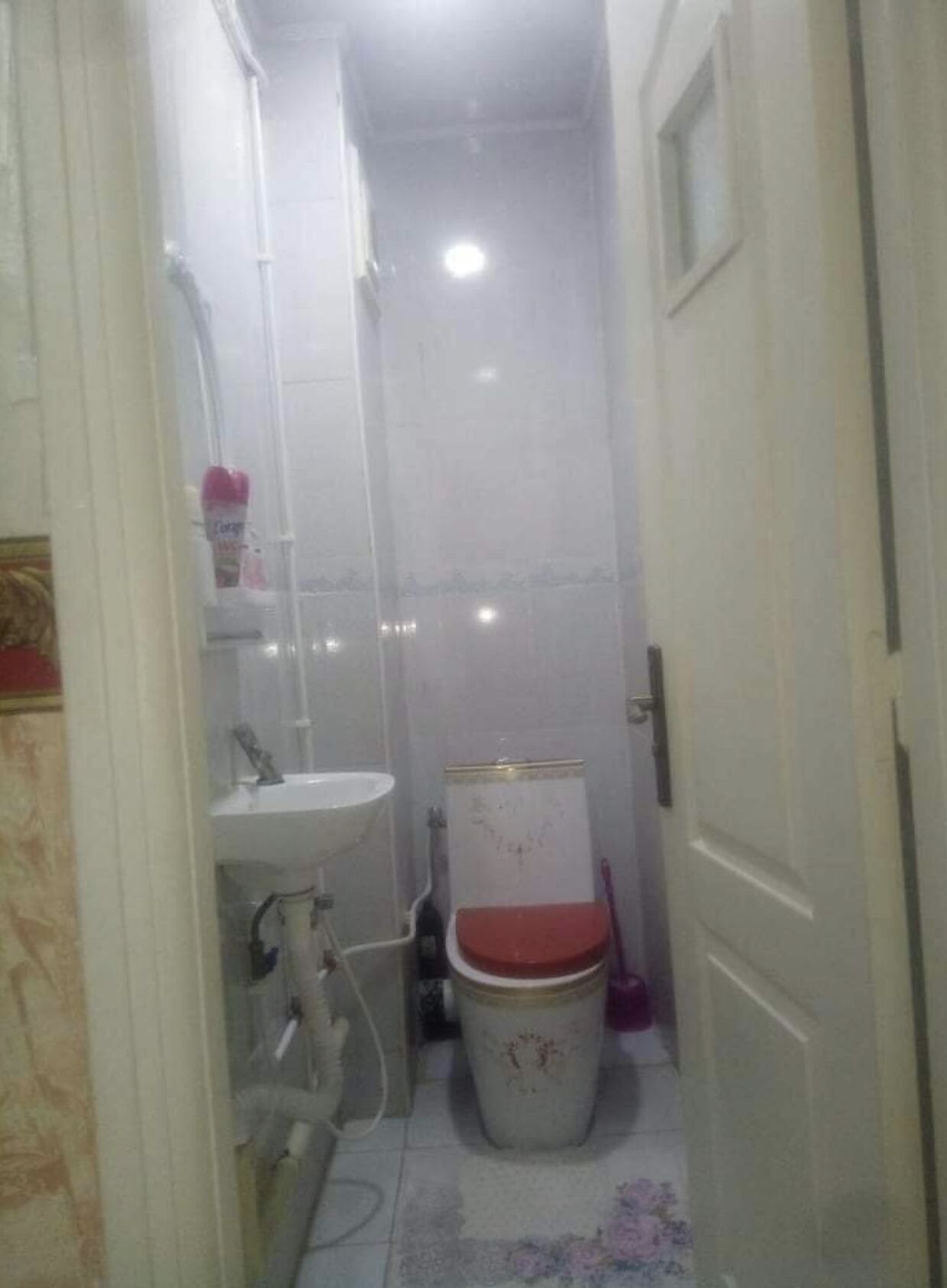 A narrow bathroom with a white sink, a toilet with a red seat, and a shower area.