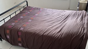 A single bed with a brown bedspread in a room with white walls, a window, and a radiator. - Chambre Chez Lhabitant (Cholet)