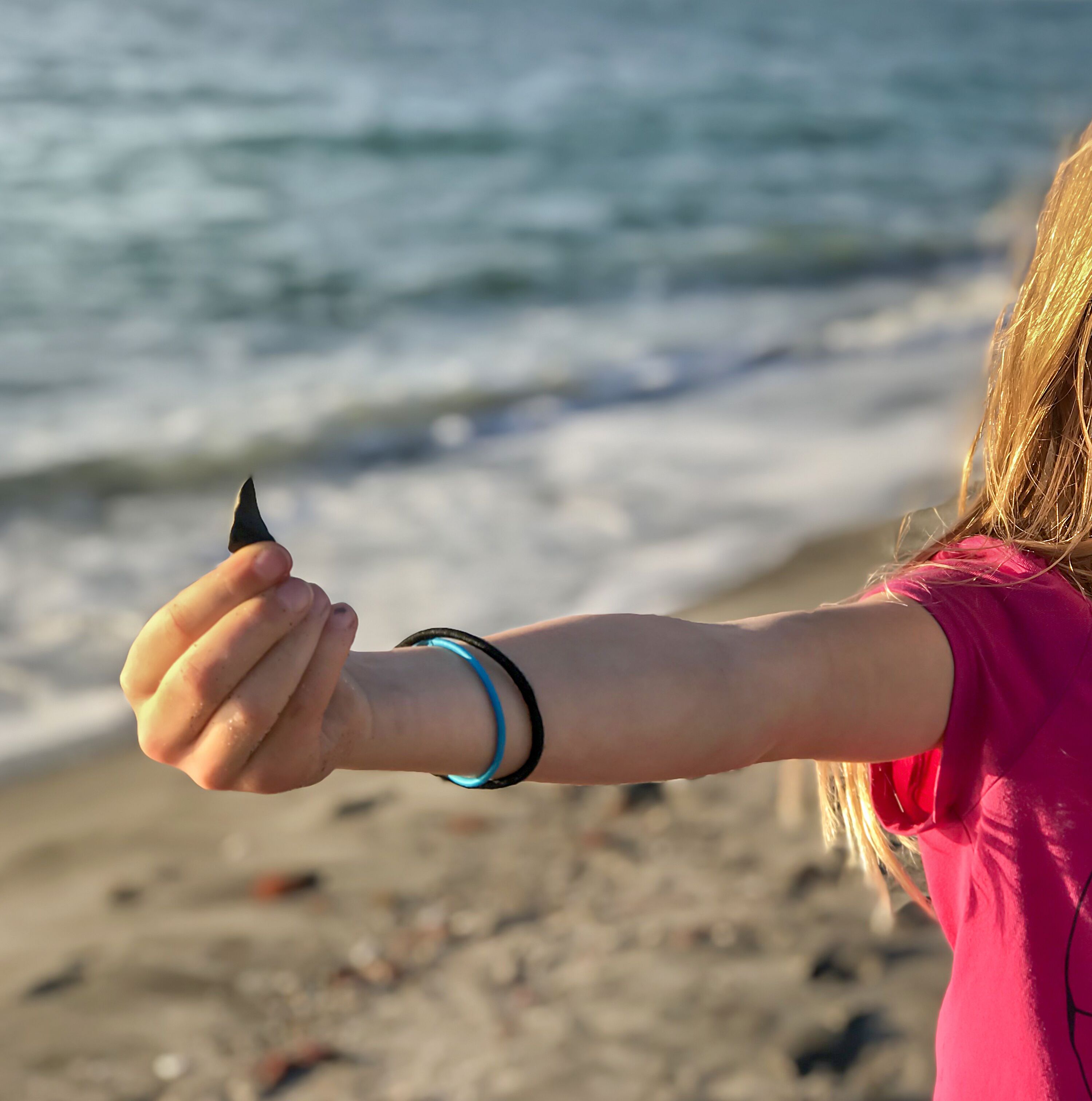 A person holding a black object by their wrist, with a beach and ocean in the background.
