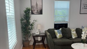 A living room with a green sofa, a small table with a lamp, a potted plant, and a cityscape artwork on the wall. - Charming Townhouse 12 minutes to Boston (Watertown)
