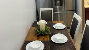 A dining area with a wooden table set for four, surrounded by chairs. A large framed botanical illustration is on the wall. A stainless steel refrigerator and a microwave are visible in the background. - Charming Townhouse 12 minutes to Boston (Watertown)