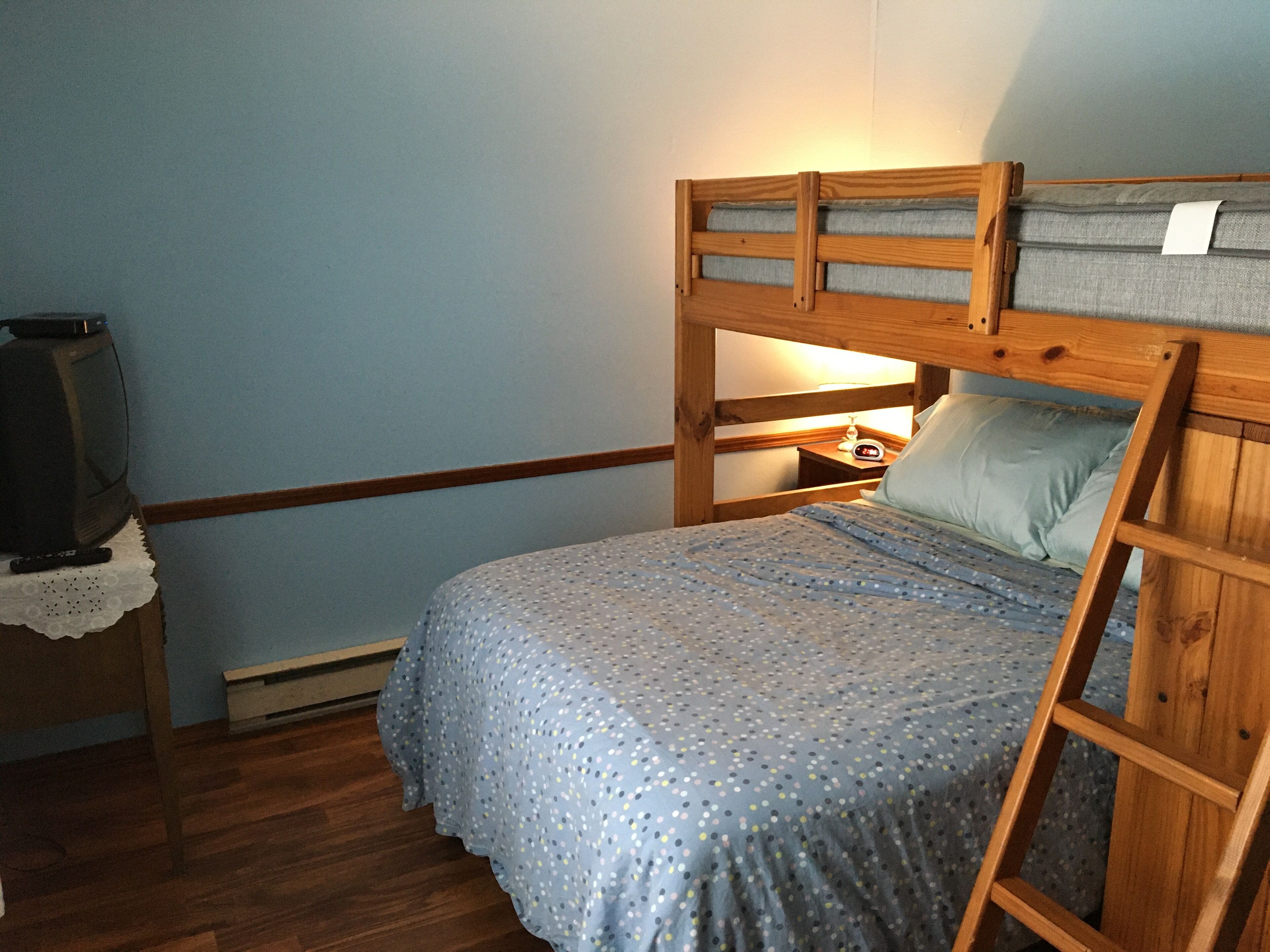 Un lit superposé avec une structure en bois et un matelas simple. Il y a une table de chevet avec une lampe et une horloge. Un téléviseur est posé sur une petite table recouverte d’une nappe en dentelle.